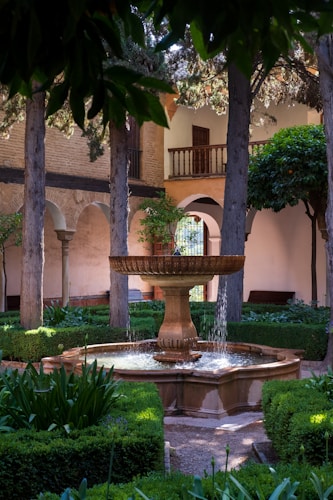 A serene garden courtyard with a stone fountain at its center, surrounded by neatly trimmed hedges and trees. The courtyard is enclosed by archways and walls with a wooden balcony visible in the background. Sunlight filters through the leaves creating patches of light and shadow on the greenery.