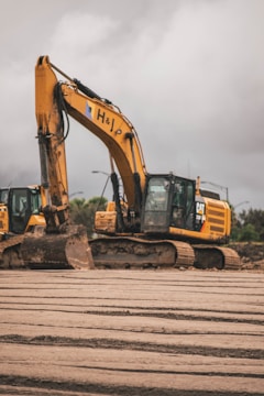 A large construction excavator sits on a muddy construction site with overcast skies in the background. The machine is yellow and has a visible logo on its side. There are tracks visible on the ground, indicating recent activity in the area. Another excavator is partially visible to the left.