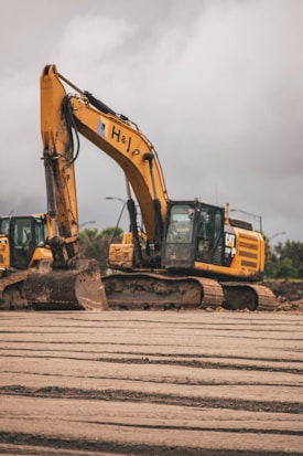 A large construction excavator sits on a muddy construction site with overcast skies in the background. The machine is yellow and has a visible logo on its side. There are tracks visible on the ground, indicating recent activity in the area. Another excavator is partially visible to the left.