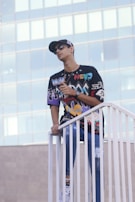 A person stands on a white railing, wearing a colorful graphic shirt, jeans, and a cap. The background consists of a modern glass building, suggesting an urban setting.