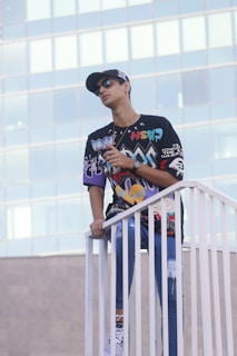 A person stands on a white railing, wearing a colorful graphic shirt, jeans, and a cap. The background consists of a modern glass building, suggesting an urban setting.