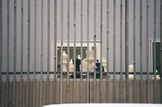 Large chess pieces are arranged on a wooden platform behind vertical metal bars. The setting appears to be outdoors against a corrugated metal wall with two small windows visible.