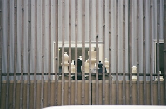 Large chess pieces are arranged on a wooden platform behind vertical metal bars. The setting appears to be outdoors against a corrugated metal wall with two small windows visible.