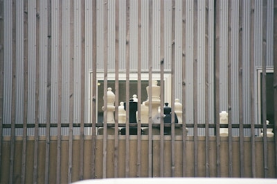 Large chess pieces are arranged on a wooden platform behind vertical metal bars. The setting appears to be outdoors against a corrugated metal wall with two small windows visible.
