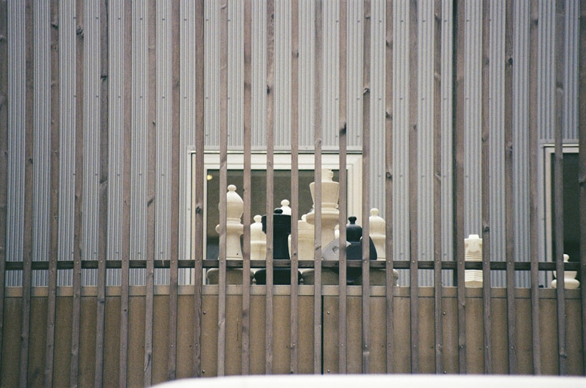 Large chess pieces are arranged on a wooden platform behind vertical metal bars. The setting appears to be outdoors against a corrugated metal wall with two small windows visible.