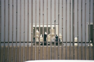 Large chess pieces are arranged on a wooden platform behind vertical metal bars. The setting appears to be outdoors against a corrugated metal wall with two small windows visible.