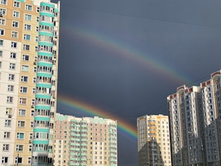 Two rainbows arch across a sky mostly covered by dark clouds, with tall residential buildings in the foreground. The buildings are multi-storey, with a combination of beige and white facades, and some feature turquoise accents on their balconies.