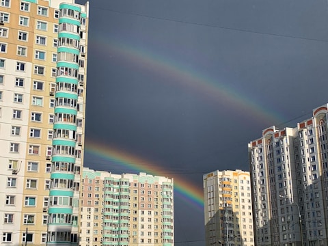 Two rainbows arch across a sky mostly covered by dark clouds, with tall residential buildings in the foreground. The buildings are multi-storey, with a combination of beige and white facades, and some feature turquoise accents on their balconies.