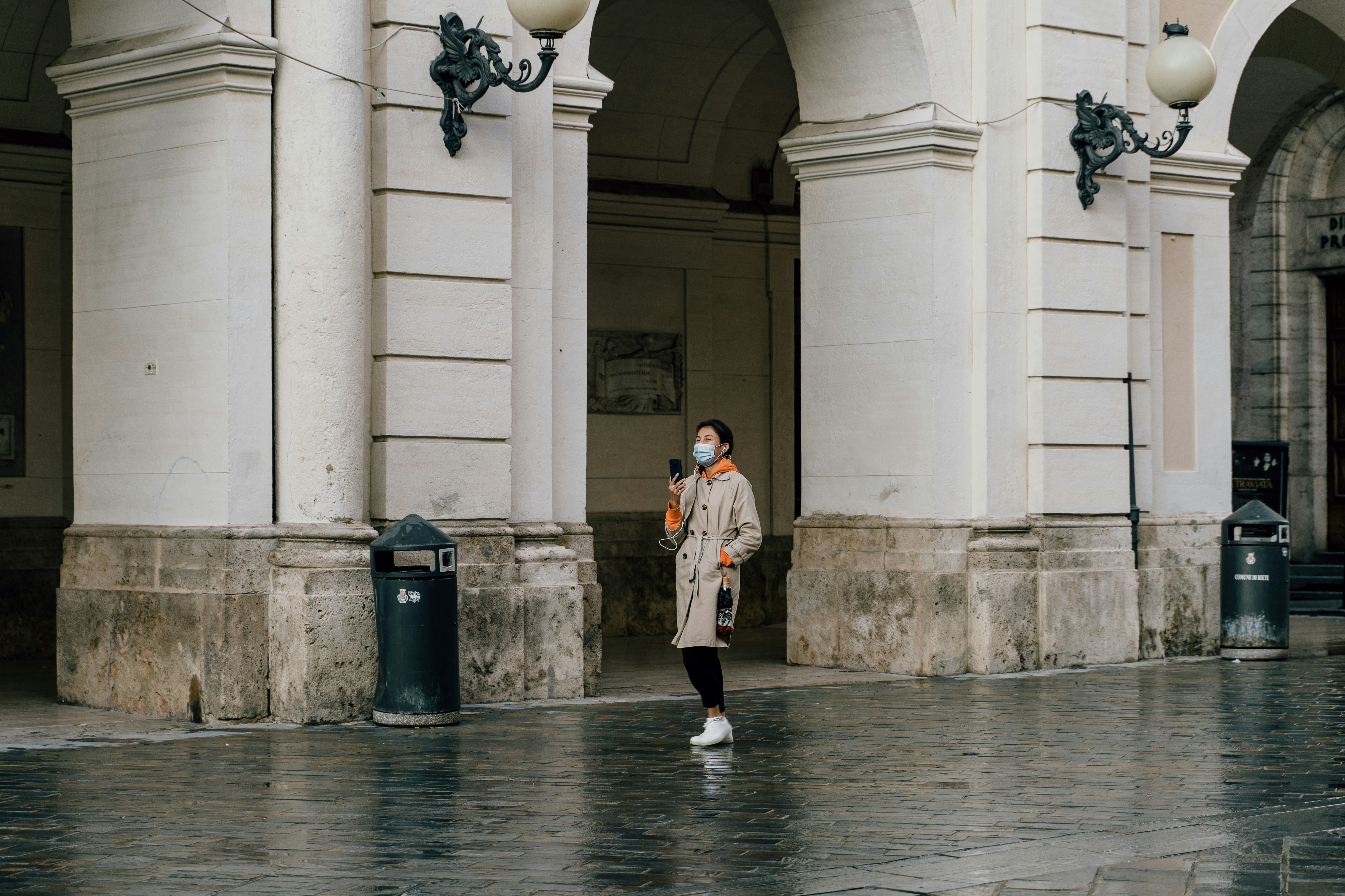 A girl, wearing a face mask, strolls across a piazza deep in conversation holding her cell phone up