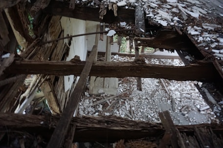 Decaying wooden beams and rubble form the remnants of a collapsed roof, with splintered wood and white debris scattered across the floor, suggesting an abandoned or neglected structure. Light filters through gaps, highlighting the aged, weathered textures.