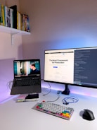A modern home office setup featuring a laptop on a stand with a video playing, a large monitor displaying a webpage about the React Framework, a colorful mechanical keyboard, and a computer mouse on a white desk. Books are neatly arranged on a shelf attached to the wall.