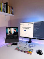 A modern home office setup featuring a laptop on a stand with a video playing, a large monitor displaying a webpage about the React Framework, a colorful mechanical keyboard, and a computer mouse on a white desk. Books are neatly arranged on a shelf attached to the wall.