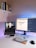 A modern home office setup featuring a laptop on a stand with a video playing, a large monitor displaying a webpage about the React Framework, a colorful mechanical keyboard, and a computer mouse on a white desk. Books are neatly arranged on a shelf attached to the wall.