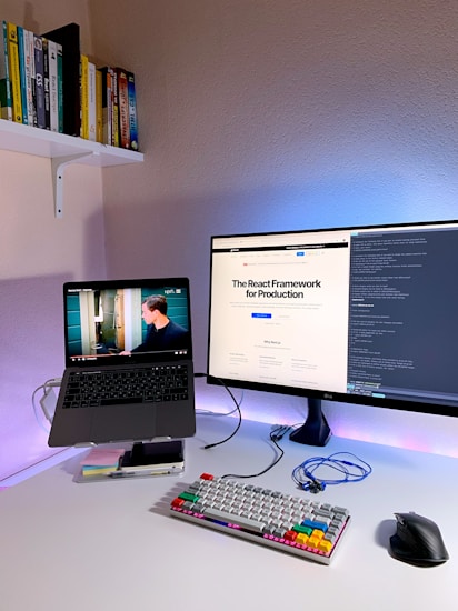 A modern home office setup featuring a laptop on a stand with a video playing, a large monitor displaying a webpage about the React Framework, a colorful mechanical keyboard, and a computer mouse on a white desk. Books are neatly arranged on a shelf attached to the wall.