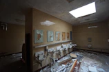 An abandoned and deteriorating restroom with broken mirrors and damaged sinks. The floor is littered with debris, including broken tiles and pipes. The walls are tiled and stained, and there is a large skylight providing some illumination.