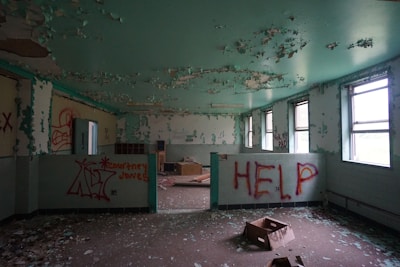 An abandoned room with peeling paint on the walls and ceiling, scattered debris, and broken furniture. Graffiti, such as the word 'HELP' and various tags, is visible on the walls. The lighting is dim, coming from overcast windows, enhancing the sense of neglect. The walls and ceiling are a faint turquoise, contrasting with the dark floor.