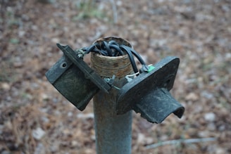 A broken electrical junction box sits atop a metal post, with exposed and tangled wires emerging from the top. The surrounding background features a blurred, earthy terrain.