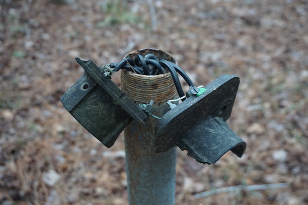 A broken electrical junction box sits atop a metal post, with exposed and tangled wires emerging from the top. The surrounding background features a blurred, earthy terrain.