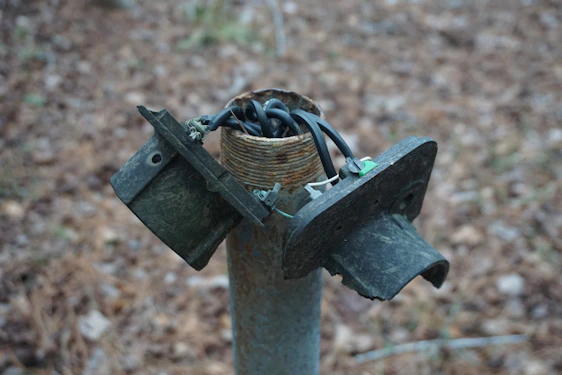 A broken electrical junction box sits atop a metal post, with exposed and tangled wires emerging from the top. The surrounding background features a blurred, earthy terrain.