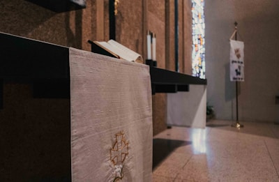 An indoor space featuring a religious setting with a white altar cloth displaying a cross emblem, an open book on the altar, and a stained glass window in the background. A banner with inscriptions and symbols stands on a pole nearby.