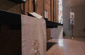 An indoor space featuring a religious setting with a white altar cloth displaying a cross emblem, an open book on the altar, and a stained glass window in the background. A banner with inscriptions and symbols stands on a pole nearby.