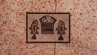 A mosaic artwork set in a floor features a design of two stylized fish flanking a basket containing what appear to be bread loaves. The mosaic is composed of small, colored tiles arranged to create the image, bordered by a thin frame. The surrounding floor consists of a terrazzo-style pattern with small speckles of red, brown, and beige.
