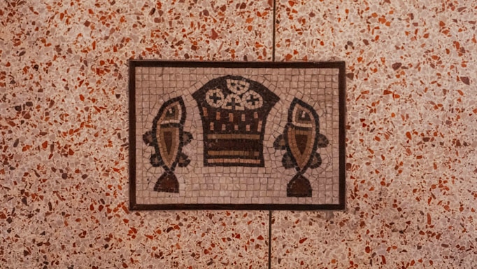 A mosaic artwork set in a floor features a design of two stylized fish flanking a basket containing what appear to be bread loaves. The mosaic is composed of small, colored tiles arranged to create the image, bordered by a thin frame. The surrounding floor consists of a terrazzo-style pattern with small speckles of red, brown, and beige.
