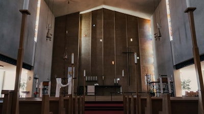 A spacious church interior with wooden pews leading towards the altar. There are tall walls with modern design and a large cross hanging at the center. Soft light filters through narrow stained glass windows on the sides, creating patches of colored illumination. The altar area is minimally decorated with religious symbols, and the atmosphere is serene and contemplative.