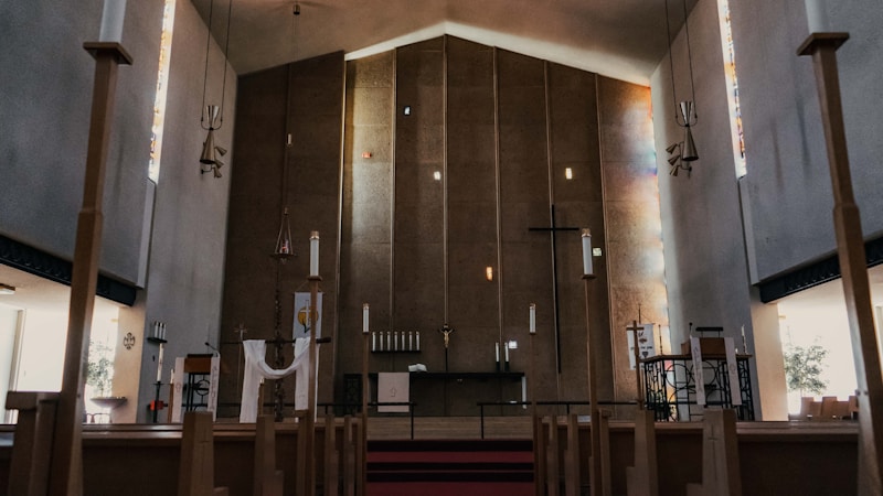 A spacious church interior with wooden pews leading towards the altar. There are tall walls with modern design and a large cross hanging at the center. Soft light filters through narrow stained glass windows on the sides, creating patches of colored illumination. The altar area is minimally decorated with religious symbols, and the atmosphere is serene and contemplative.