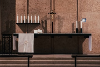 An altar with a crucifix and several candles is seen against a textured wall. The altar also holds an open book and a white cloth with an embroidered design. Shadows of the candles create an atmospheric effect.