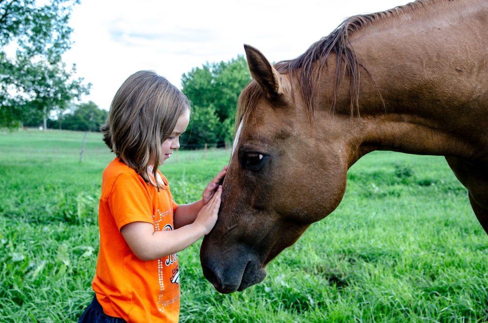 girl in orange t-shirt standing beside brown horse during daytime