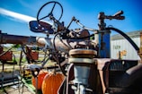 A rusty, vintage piece of farming machinery is prominently visible. The machinery is equipped with a steering wheel and metal components showing signs of wear and rust. An orange pumpkin is placed on part of the machinery, and in the background, there is a building with the word 'FARM' painted on it alongside a pumpkin illustration. The sky is clear and blue, indicating a bright day.