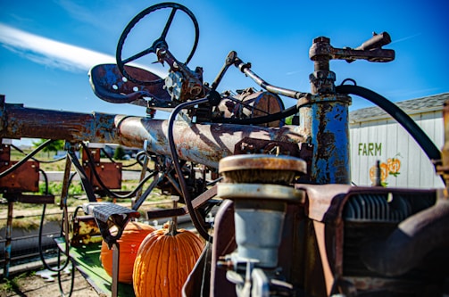 A rusty, vintage piece of farming machinery is prominently visible. The machinery is equipped with a steering wheel and metal components showing signs of wear and rust. An orange pumpkin is placed on part of the machinery, and in the background, there is a building with the word 'FARM' painted on it alongside a pumpkin illustration. The sky is clear and blue, indicating a bright day.