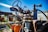 A rusty, vintage piece of farming machinery is prominently visible. The machinery is equipped with a steering wheel and metal components showing signs of wear and rust. An orange pumpkin is placed on part of the machinery, and in the background, there is a building with the word 'FARM' painted on it alongside a pumpkin illustration. The sky is clear and blue, indicating a bright day.