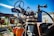 A rusty, vintage piece of farming machinery is prominently visible. The machinery is equipped with a steering wheel and metal components showing signs of wear and rust. An orange pumpkin is placed on part of the machinery, and in the background, there is a building with the word 'FARM' painted on it alongside a pumpkin illustration. The sky is clear and blue, indicating a bright day.