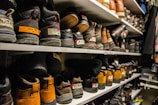 Shelves filled with rows of various pairs of boots and shoes are seen, each with labels and tags attached for identification. The footwear appears to be organized meticulously, possibly indicating a storage area or inventory space. The room lighting creates a warm tone, highlighting the textures and materials of the shoes.
