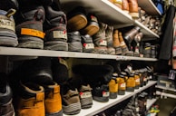 Shelves filled with rows of various pairs of boots and shoes are seen, each with labels and tags attached for identification. The footwear appears to be organized meticulously, possibly indicating a storage area or inventory space. The room lighting creates a warm tone, highlighting the textures and materials of the shoes.