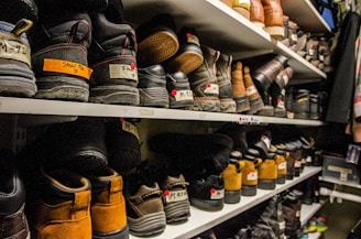Shelves filled with rows of various pairs of boots and shoes are seen, each with labels and tags attached for identification. The footwear appears to be organized meticulously, possibly indicating a storage area or inventory space. The room lighting creates a warm tone, highlighting the textures and materials of the shoes.
