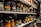 Shelves filled with rows of various pairs of boots and shoes are seen, each with labels and tags attached for identification. The footwear appears to be organized meticulously, possibly indicating a storage area or inventory space. The room lighting creates a warm tone, highlighting the textures and materials of the shoes.