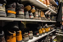 Shelves filled with rows of various pairs of boots and shoes are seen, each with labels and tags attached for identification. The footwear appears to be organized meticulously, possibly indicating a storage area or inventory space. The room lighting creates a warm tone, highlighting the textures and materials of the shoes.