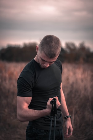 A person wearing a black t-shirt is standing outdoors, looking down while holding an exercise resistance band. The background consists of tall grass fields under an overcast sky, creating a serene and focused atmosphere.
