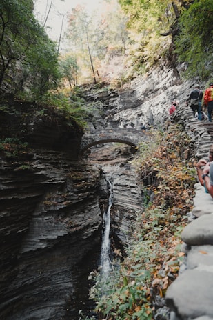 Hikers crossing a narrow stone bridge over a sparkling mountain stream surrounded by lush greenery.
