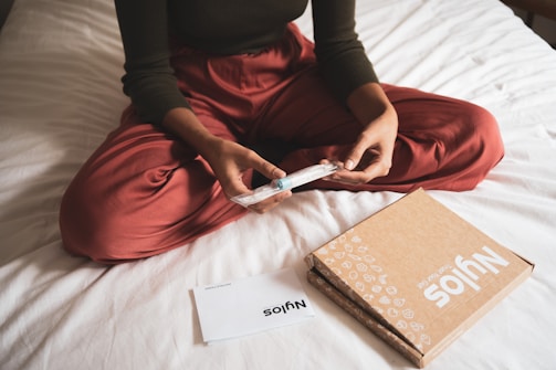 A person in red pants is sitting cross-legged on a bed, holding a packaged item. Nearby, there is a cardboard box with branding and a folded paper. The setting appears relaxed and casual.