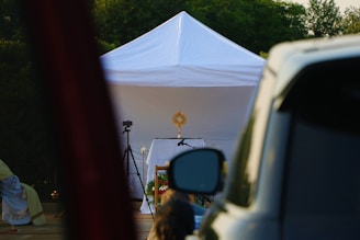 A white tent is set up outdoors with a religious monstrance displayed on a table inside. A camera on a tripod is positioned nearby, and a vehicle is partially visible in the foreground. Greenery is visible in the background.