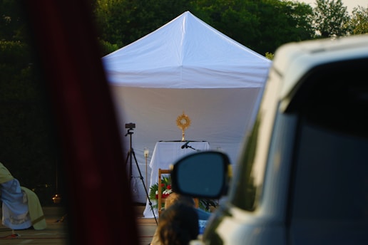 A white tent is set up outdoors with a religious monstrance displayed on a table inside. A camera on a tripod is positioned nearby, and a vehicle is partially visible in the foreground. Greenery is visible in the background.