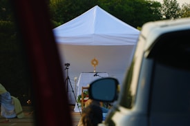 A white tent is set up outdoors with a religious monstrance displayed on a table inside. A camera on a tripod is positioned nearby, and a vehicle is partially visible in the foreground. Greenery is visible in the background.