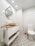 A modern bathroom featuring white subway tiles on the walls and a stylish patterned floor. A round, slightly tilted, mirror hangs above a sleek vanity with a wooden countertop and a white basin. The bathroom is well-lit with recessed ceiling lights. Next to the vanity is a toilet with a gold-toned toilet paper holder.