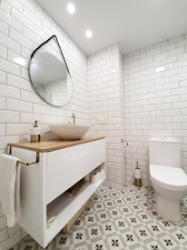 A modern bathroom featuring white subway tiles on the walls and a stylish patterned floor. A round, slightly tilted, mirror hangs above a sleek vanity with a wooden countertop and a white basin. The bathroom is well-lit with recessed ceiling lights. Next to the vanity is a toilet with a gold-toned toilet paper holder.