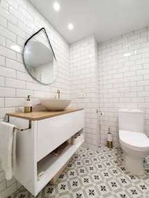 A modern bathroom featuring white subway tiles on the walls and a stylish patterned floor. A round, slightly tilted, mirror hangs above a sleek vanity with a wooden countertop and a white basin. The bathroom is well-lit with recessed ceiling lights. Next to the vanity is a toilet with a gold-toned toilet paper holder.