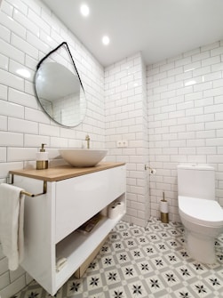 A modern bathroom featuring white subway tiles on the walls and a stylish patterned floor. A round, slightly tilted, mirror hangs above a sleek vanity with a wooden countertop and a white basin. The bathroom is well-lit with recessed ceiling lights. Next to the vanity is a toilet with a gold-toned toilet paper holder.